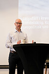 En mann står ved et svart podium med en kopp i hånden foran en projeksjonsskjerm.