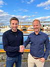 To menn håndhilser foran båter og industri ved en havn.